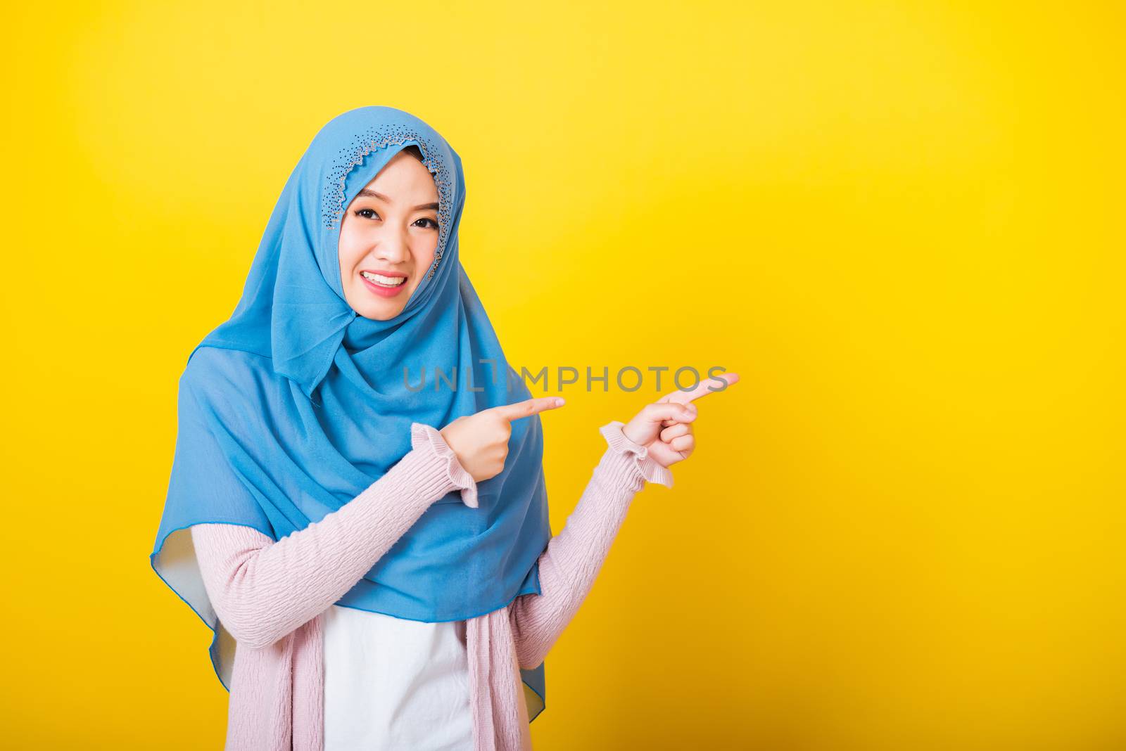 Asian Muslim Arab, Portrait of happy beautiful young woman Islam religious wear veil hijab funny smile she positive expression pointing finger side sideways to space isolated yellow background