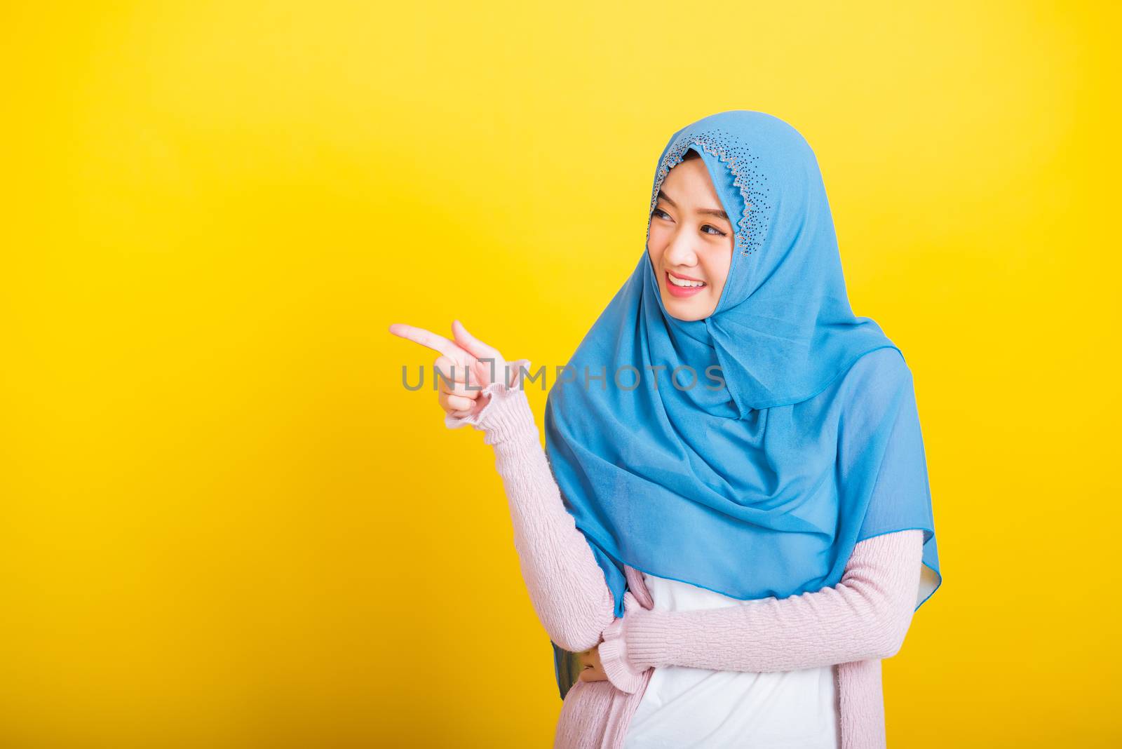 Asian Muslim Arab, Portrait of happy beautiful young woman Islam religious wear veil hijab funny smile she positive expression pointing finger side sideways to space isolated yellow background