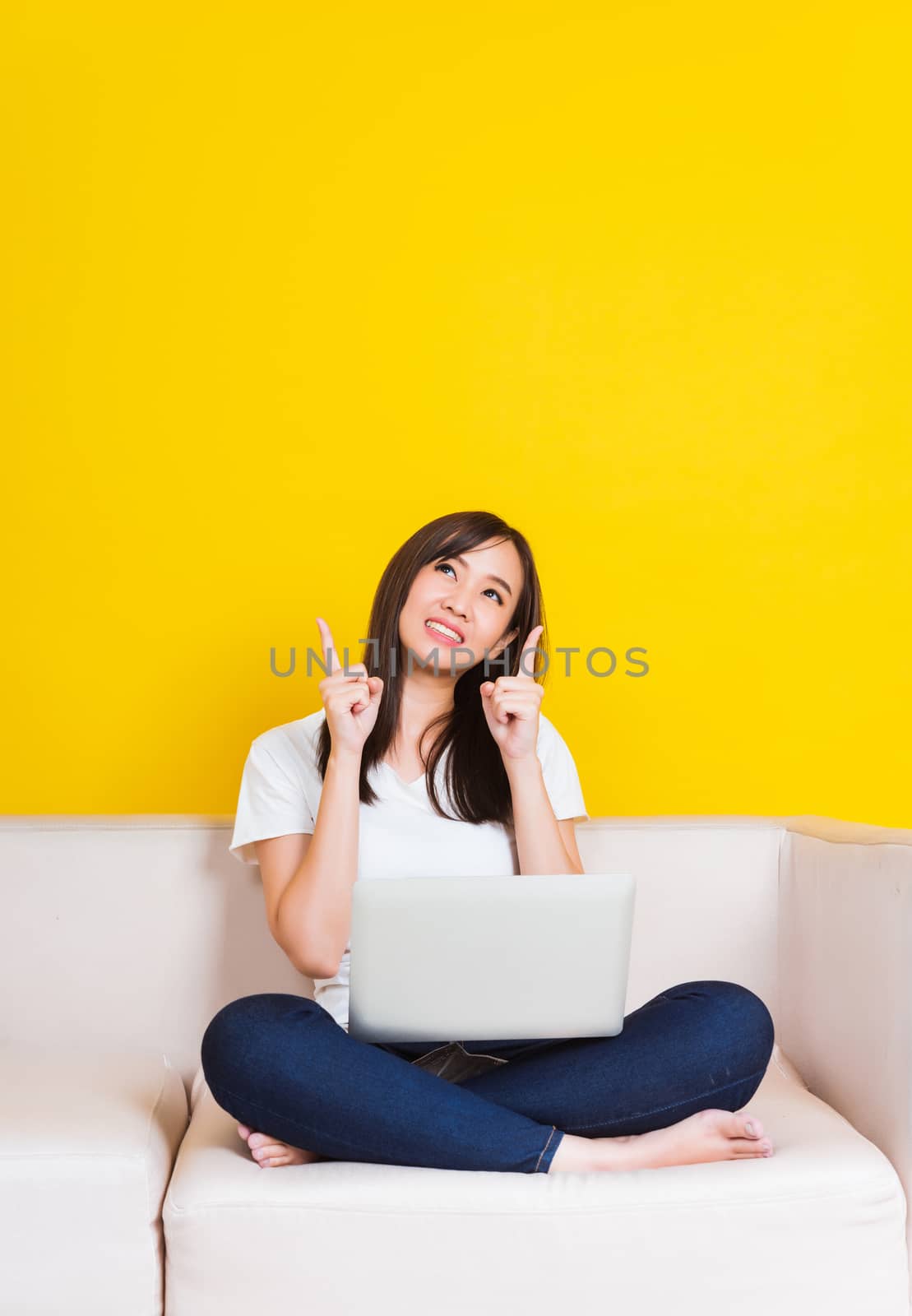 Portrait Asian of happy beautiful young woman work from home she sitting on sofa using laptop computer in house living room pointing finger up away studio shot isolated on yellow background