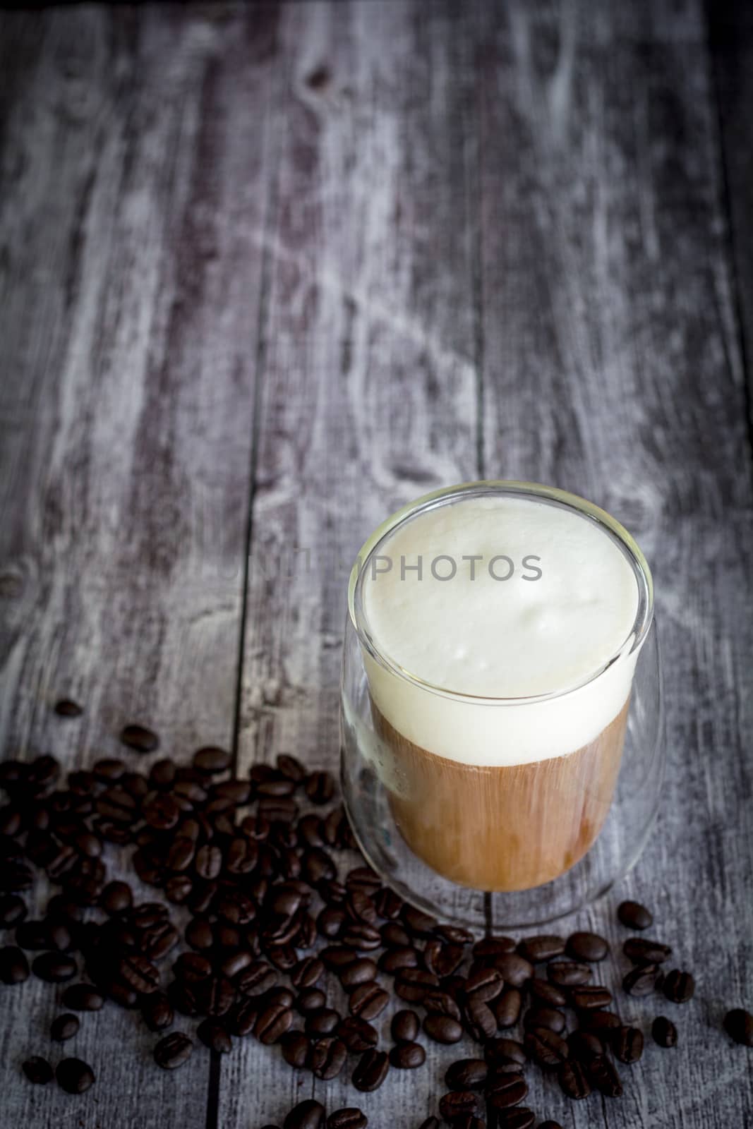 Coffee time and refreshment concept. Transparent glass of coffee with milk froth and roasted coffee bean on wooden background.