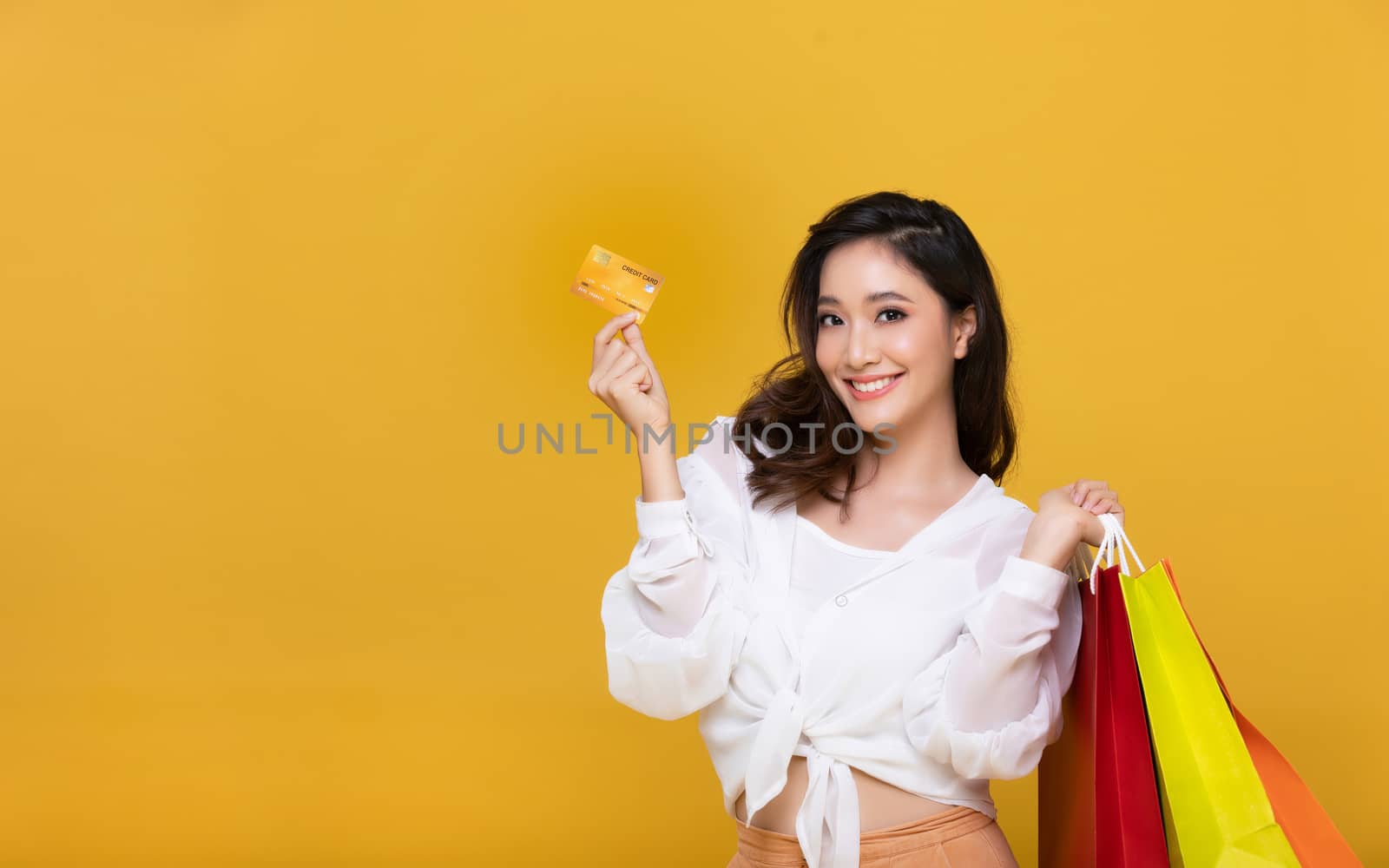 Portrait Asian beautiful happy young woman smiling cheerful and she is holding credit card and using smart phone for shopping online with shopping bags on yellow background.Happiness, consumerism, sale and people shopping concept