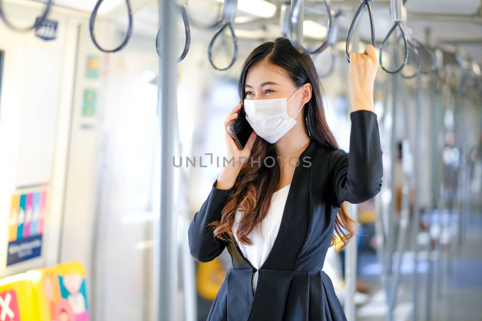 Portrait of beautiful business woman with hygiene mask use mobile phone and stand with hold handrail in sky train. She still work during pandemic of Covid-19 coronavirus spread in city.