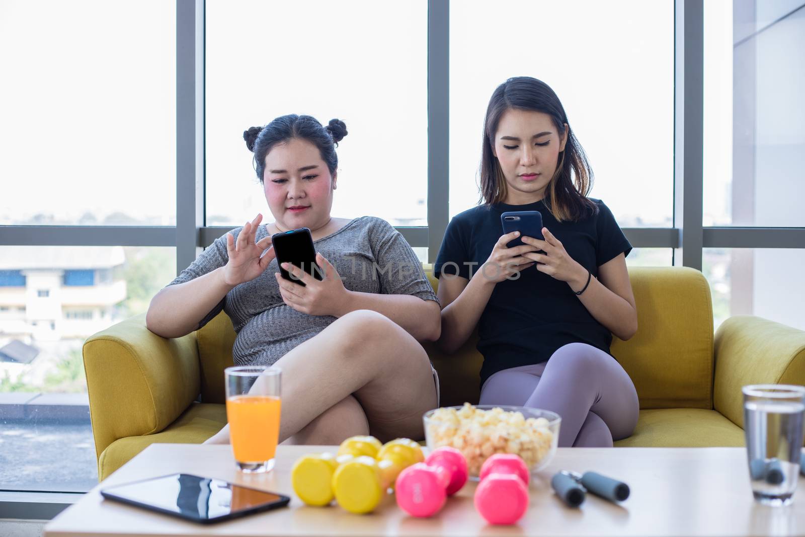 asian Overweight woman with friend play mobile phone and enjoy eating food and popcorn on sofa at home