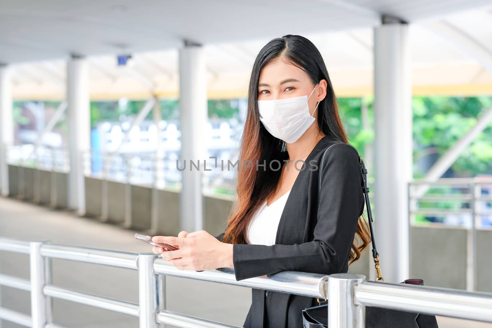 Portrait of business woman with hygiene mask stand along walk way and hold mobile phone, she also look forward and look relax during covid-19 pandemic in the city.