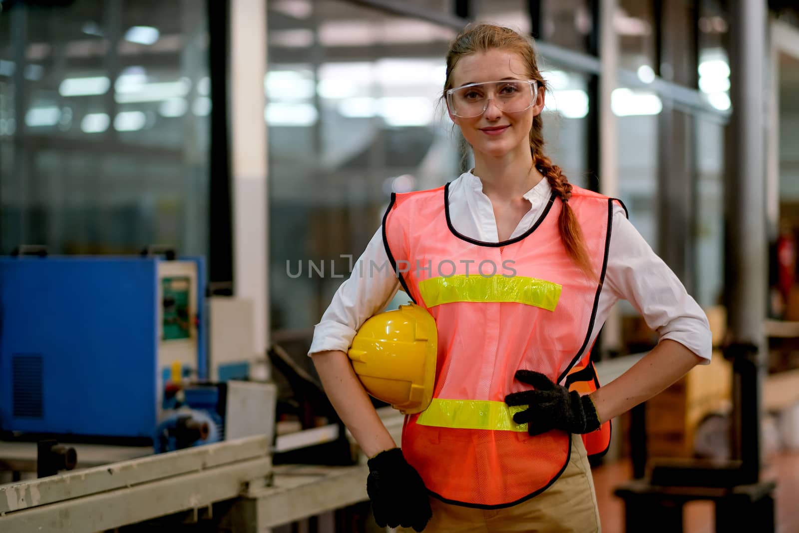 Pretty technician or engineer or worker action of akimbo and hold yellow helmet stand and smile in factory workplace.