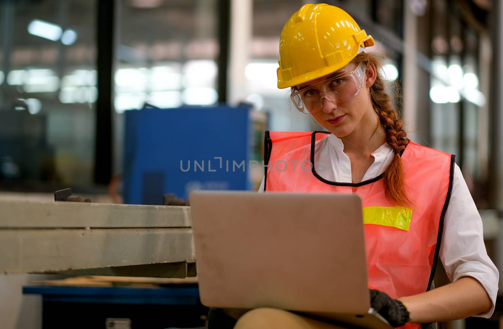 Pretty technician or engineer or worker sit on chair and use laptop in factory.