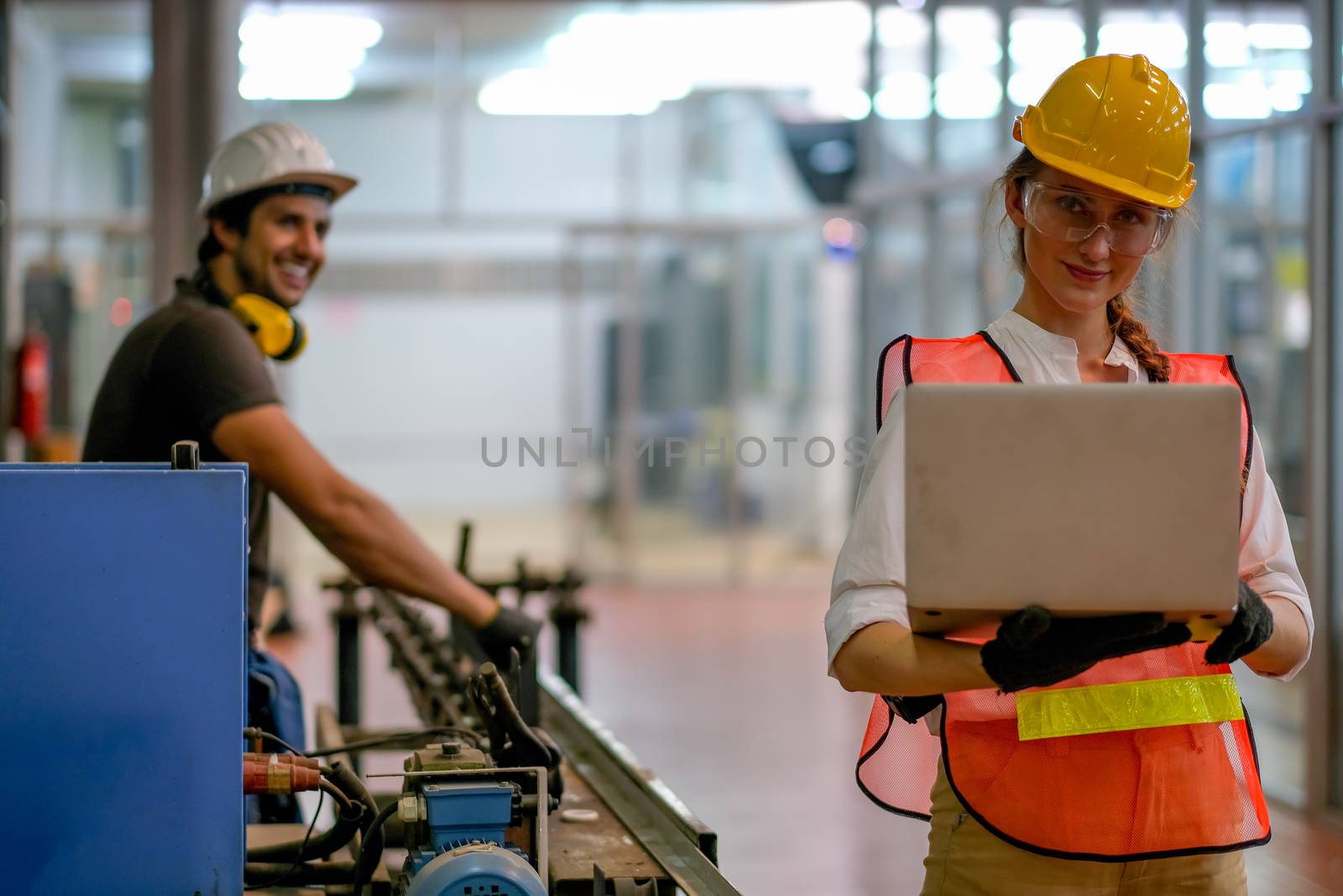 Pretty engineer or technician or worker woman hold the laptop and her co-worker stand and smile in the background and they work together in factory.