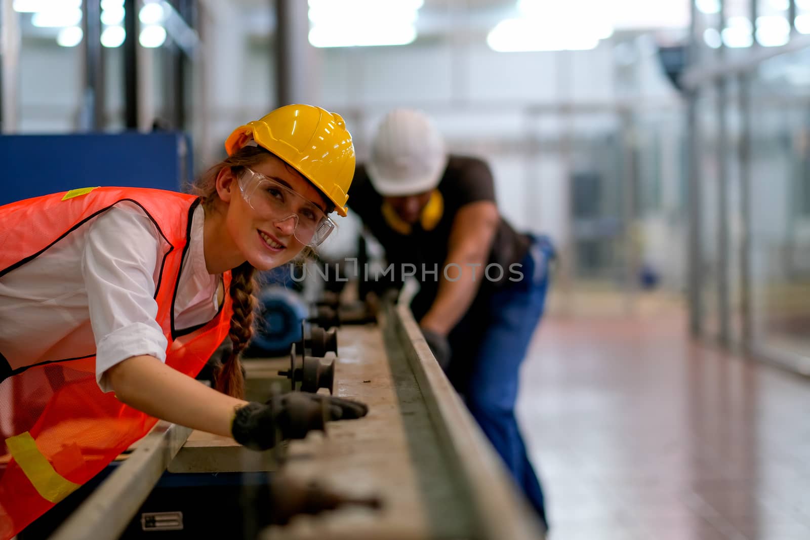 Beautiful worker or technician or engineer woman smile and look forward in front of rail of the machine with her co-worker as background in factory.