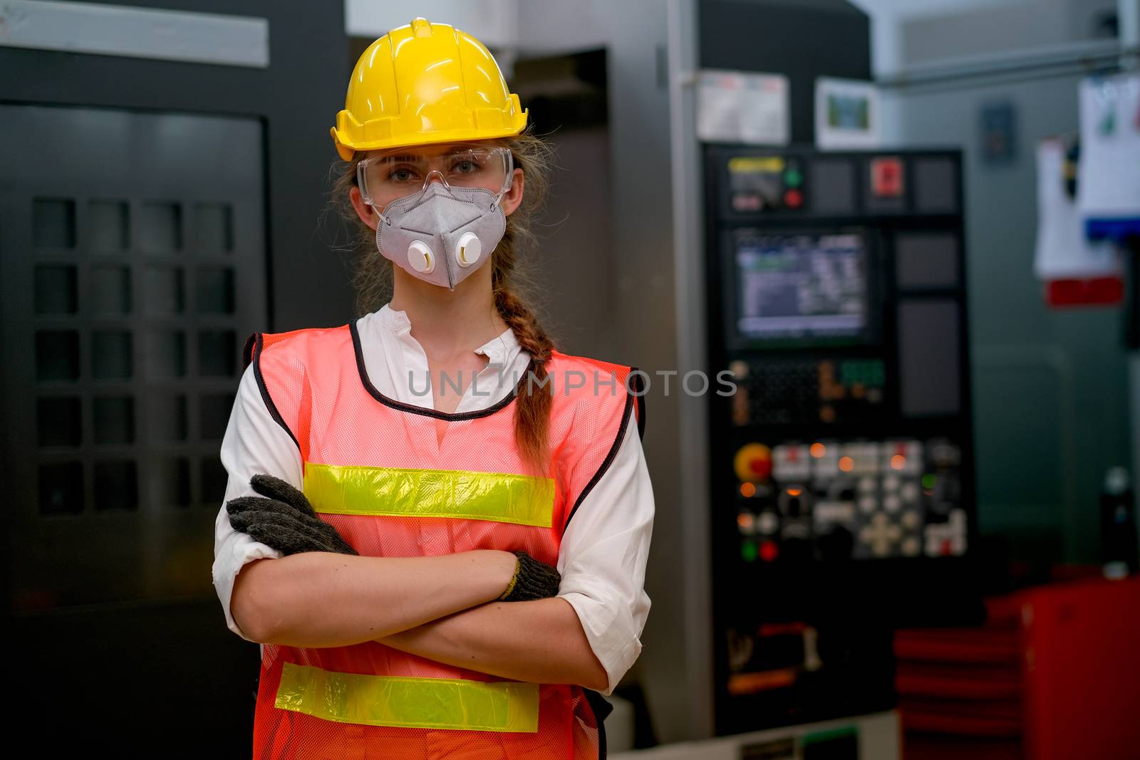 Pretty technician or worker woman with gray mask and yellow helmet stand with confident action in front of factory machine.
