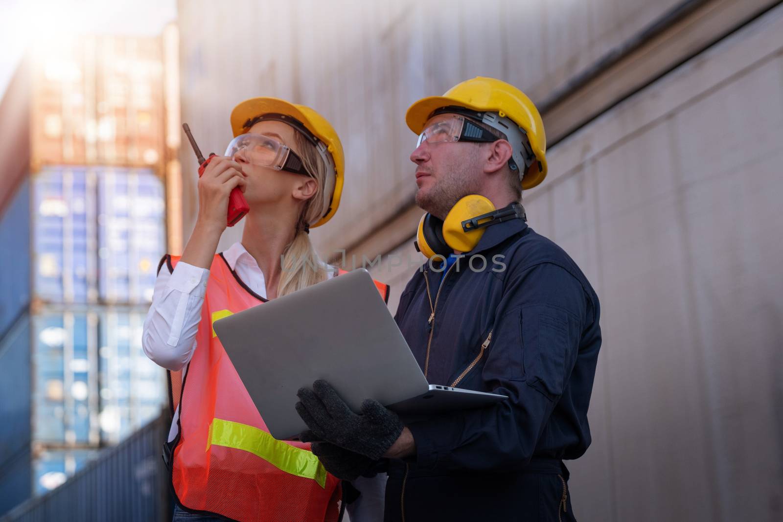 Beautiful technician or worker woman use walkie talky to communicate with her team and work with engineer man who hold laptop computer in shipping container area with day light.