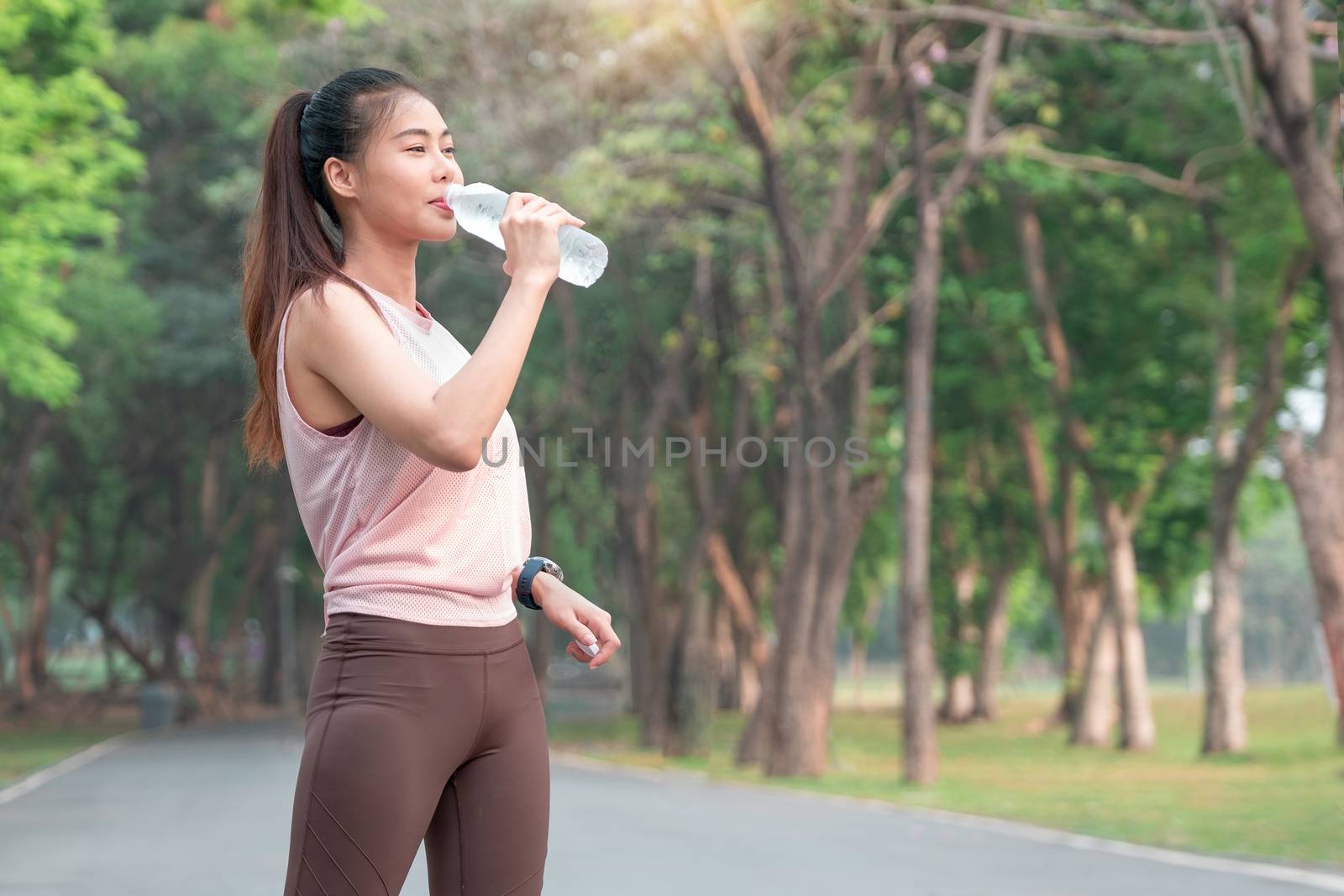 Portrait of young beautiful sport girl stand and drink bottle water after exercise with jogging in the garden or park with morning light.