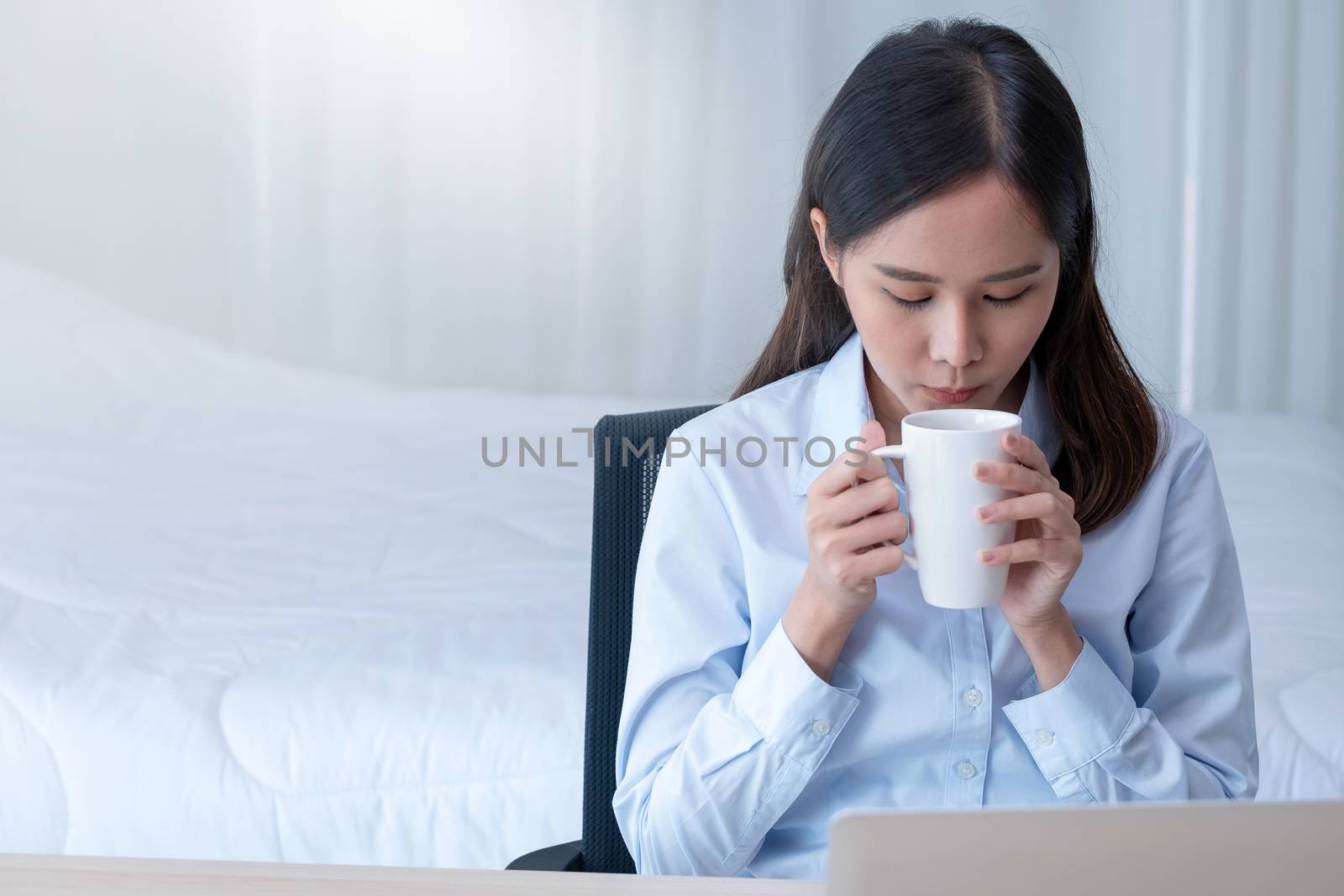Front view of pretty business girl with blue shirt drink coffee from ceramic glass during work at home in the bedroom with morning light.She look relax and happy even though corona virus pandemic.