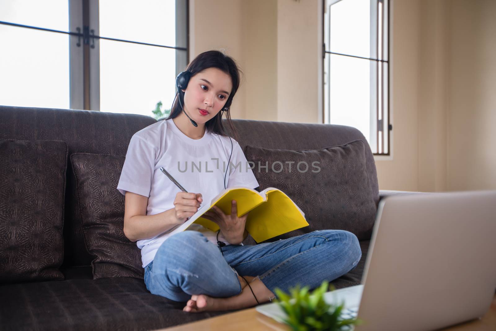 Asian happy woman student wear headphones study online watching webinar podcast on laptop listening learning education course conference calling and write notes look at laptop sit on sofa at home