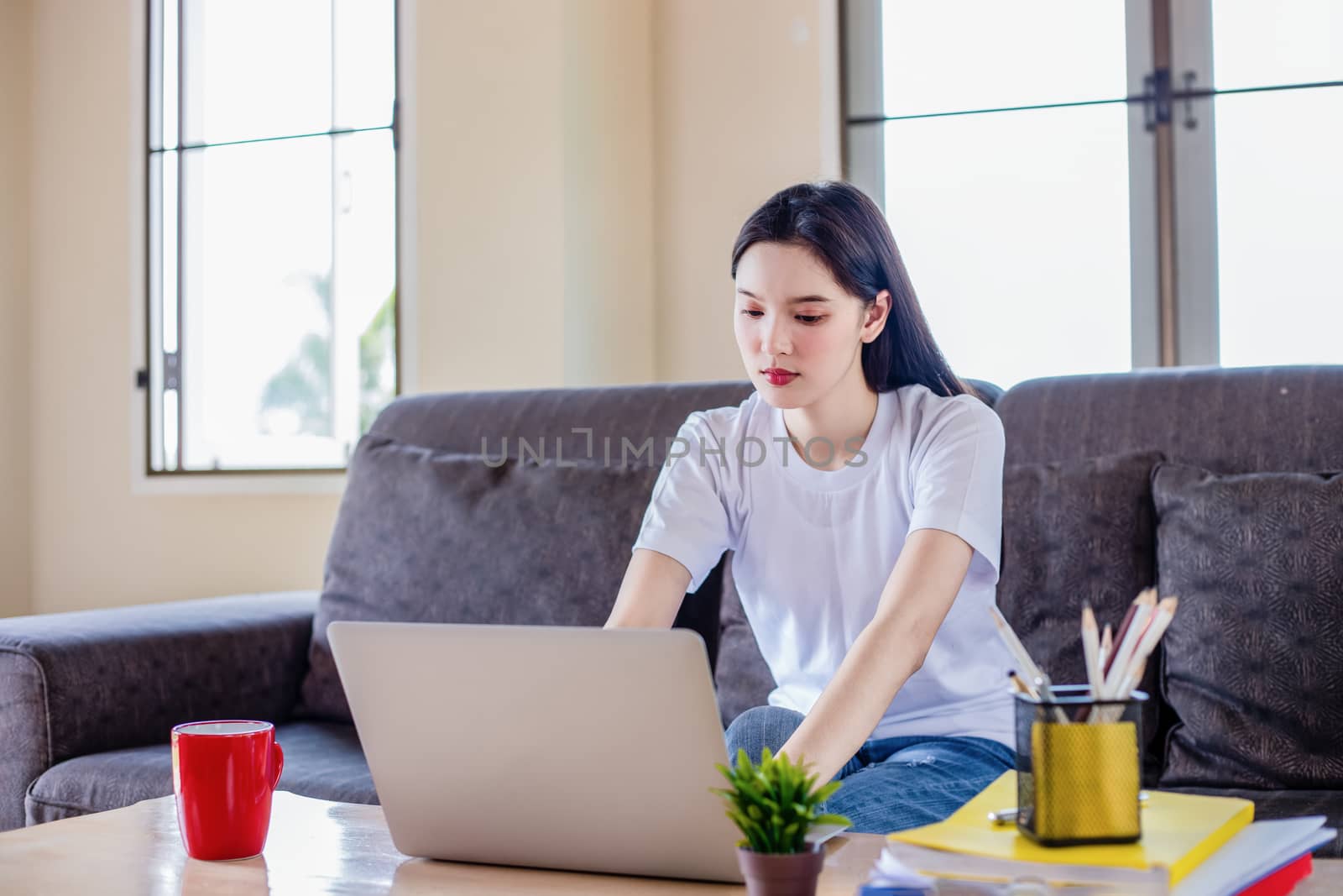 Asian woman pleased happy cheerful cute beautiful is using a notebook computer and sitting on sofa for working from home. She is quarantined at home during the coronavirus epidemic.