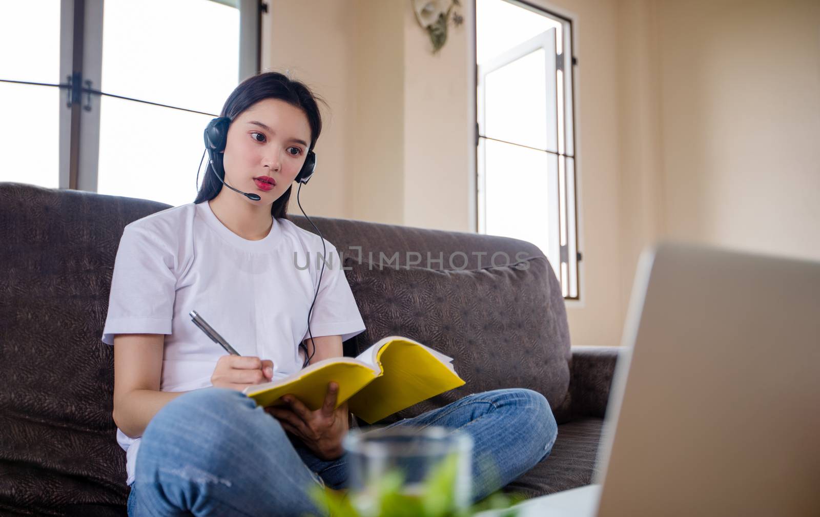 Asian happy woman student wear headphones study online watching webinar podcast on laptop listening learning education course conference calling and write notes look at laptop sit on sofa at home