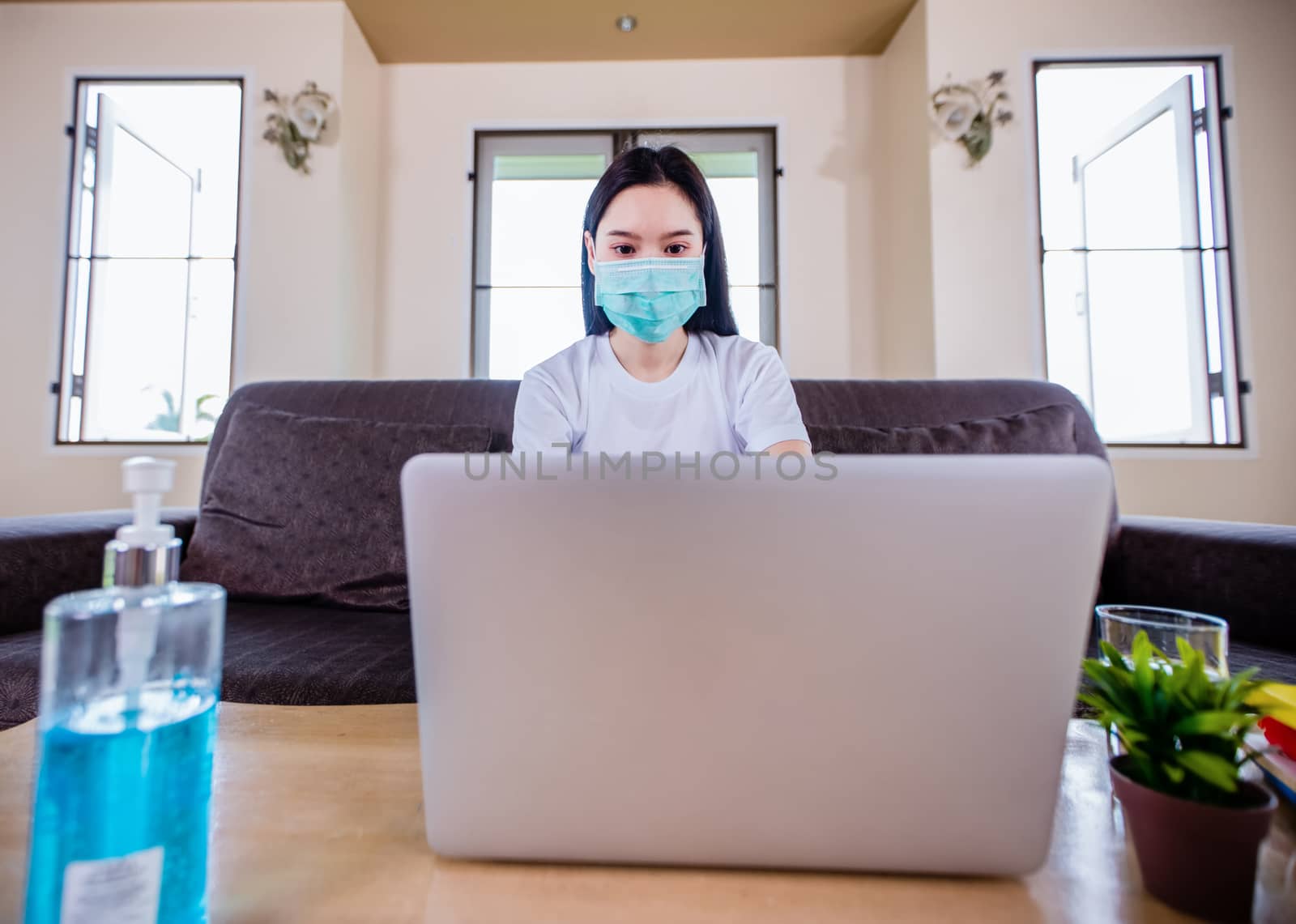Asian woman cute beautiful is wearing protective mask and using a notebook computer and sitting on sofa for working from home. She is quarantined at home .And she has sanitizer gel on the table 