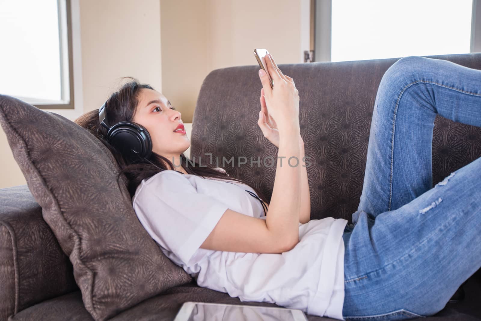 Asian young woman happy cheerful cute beautiful wears a headset for listening to music from a mobile phone and she is playing smartphone and lying on the sofa at home, 