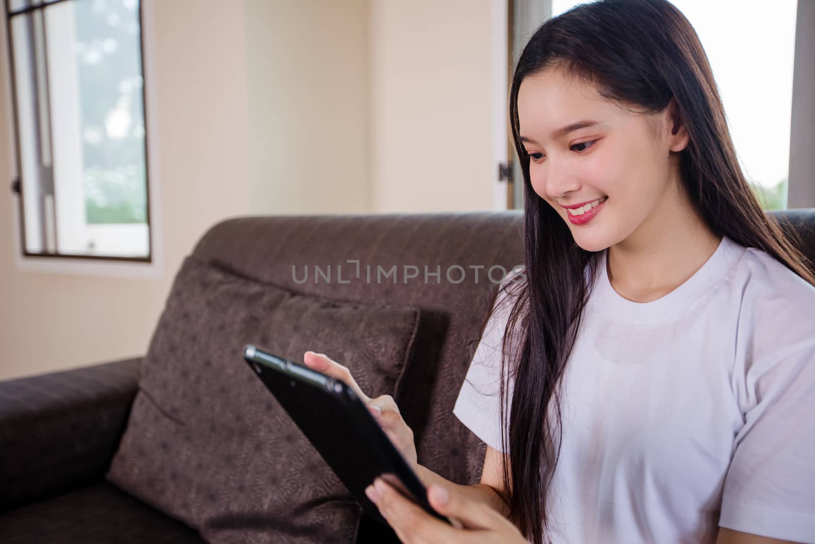 Asian young woman happy cheerful cute beautiful is playing digital pad tablet computer and sitting on the sofa at home, smiling lady using app reading electronic book relax in living room 