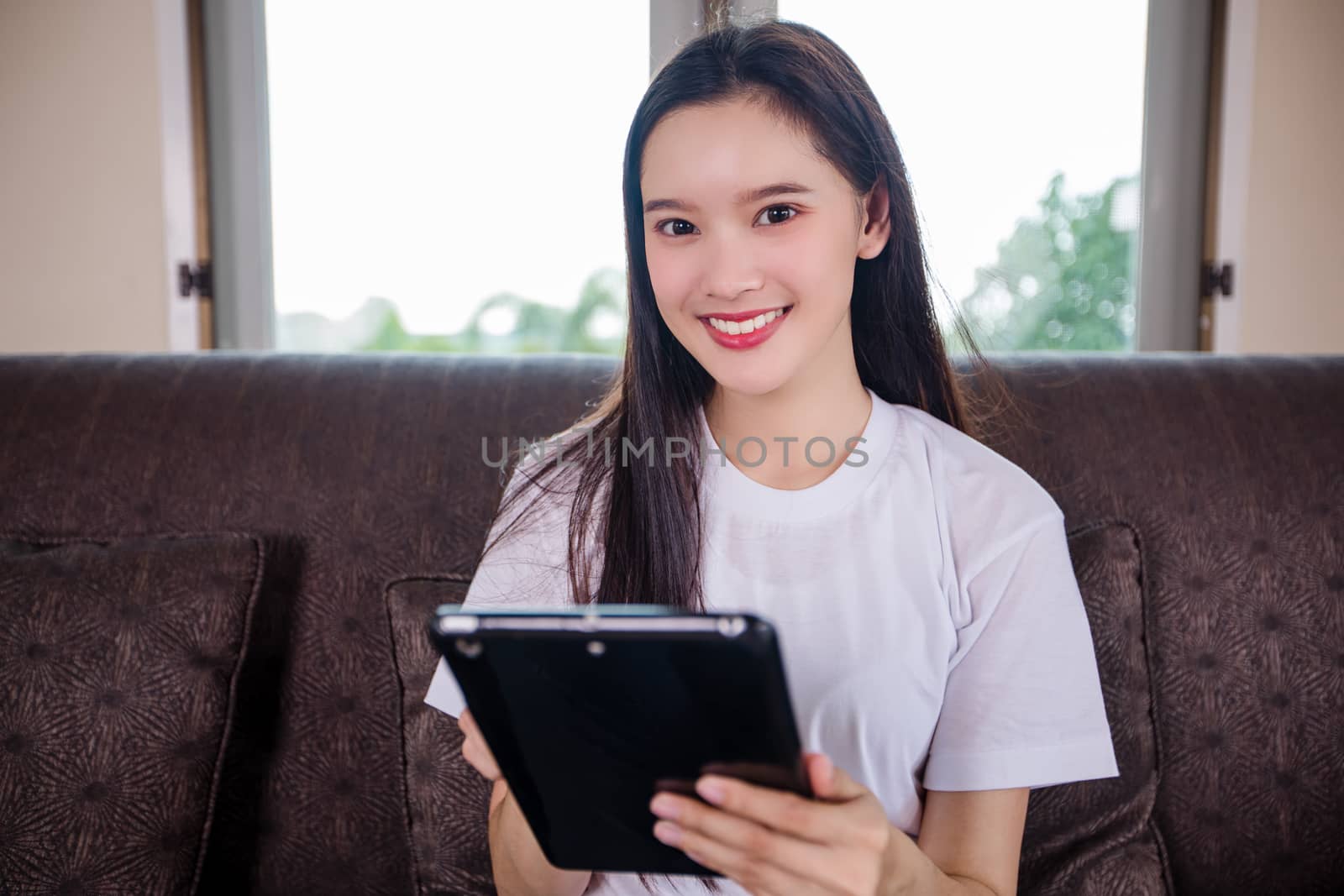 Asian young woman happy cheerful cute beautiful is playing digital pad tablet computer and sitting on the sofa at home, smiling lady using app reading electronic book relax in living room 