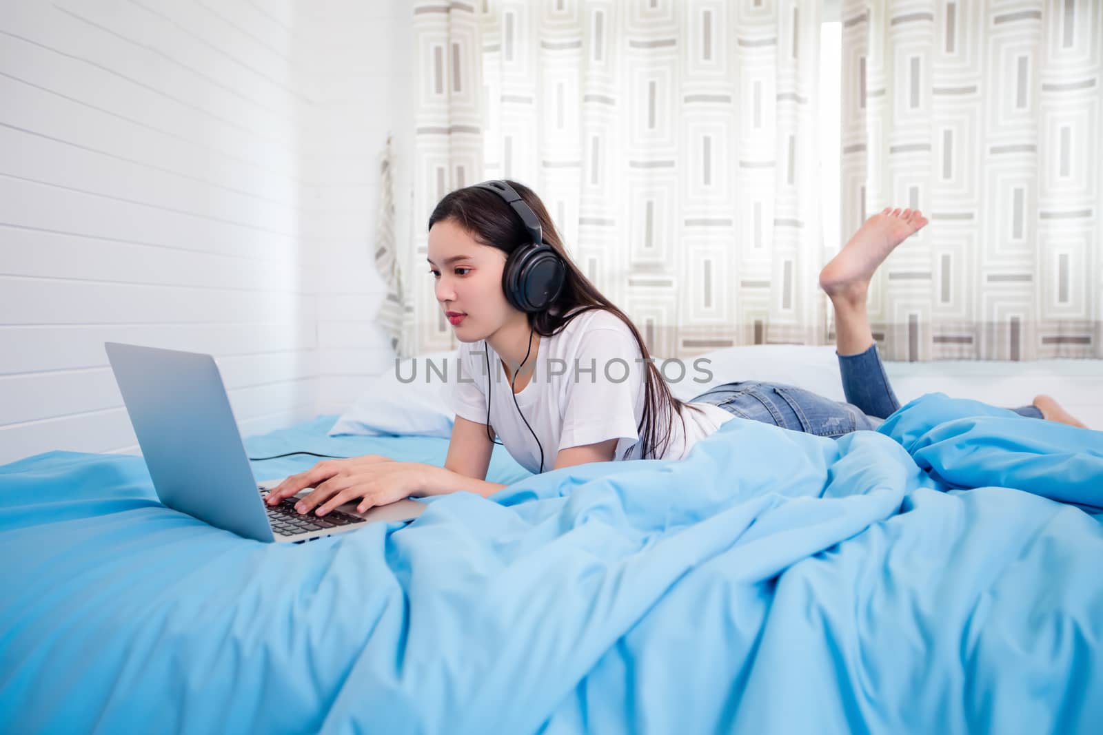 Asian happy young woman wear headphones and listening music song and watching website for shopping online from laptop  on bed at home