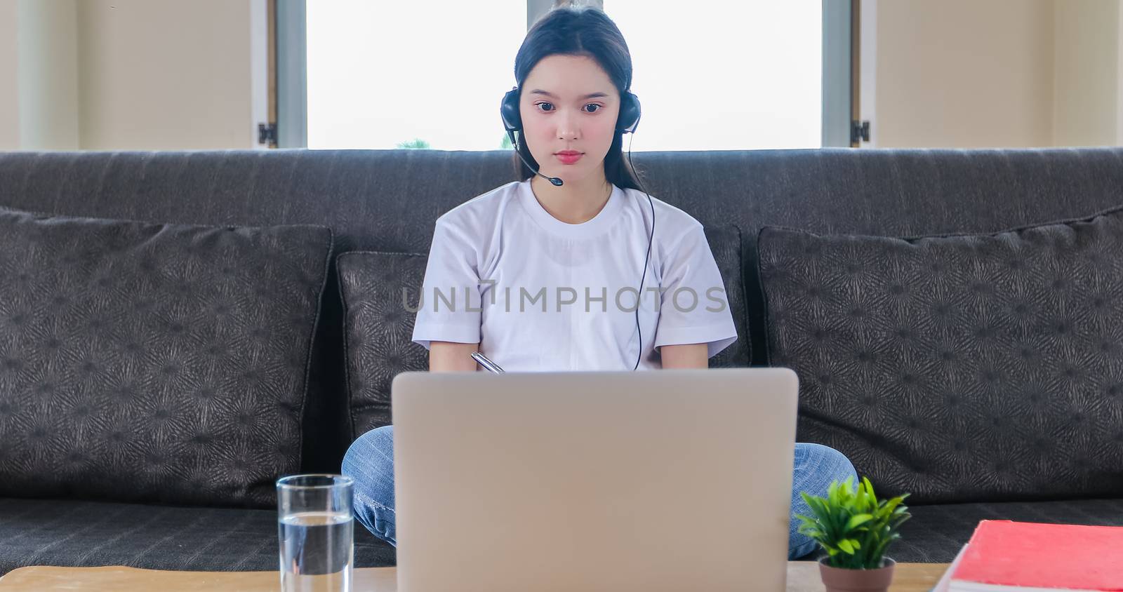 Asian happy woman student wear headphones study online watching webinar podcast on laptop listening learning education course conference calling and write notes look at laptop sit on sofa at home