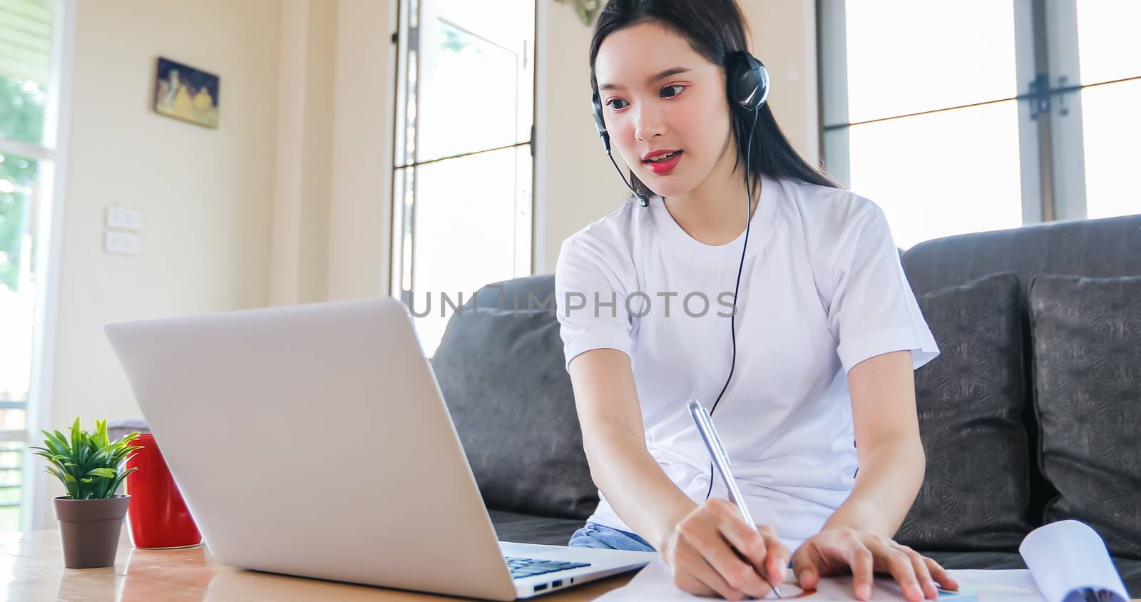 Asian happy woman student wear headphones study online watching webinar podcast on laptop listening learning education course conference calling and write notes look at laptop sit on sofa at home