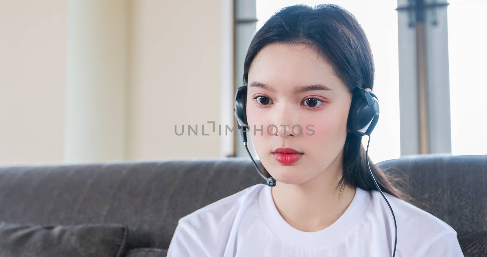 Asian happy woman student wear headphones study online watching webinar podcast on laptop listening learning education course conference calling and write notes look at laptop sit on sofa at home