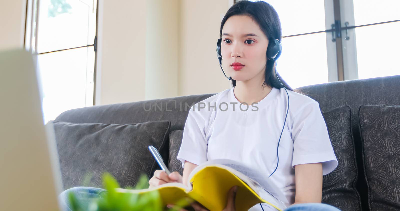 Asian happy woman student wear headphones study online watching webinar podcast on laptop listening learning education course conference calling and write notes look at laptop sit on sofa at home