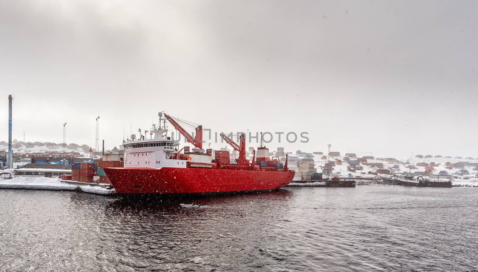 Big red cargo ship under heavy snowfall in the port of Aasiaat, with village in the background, Greenland