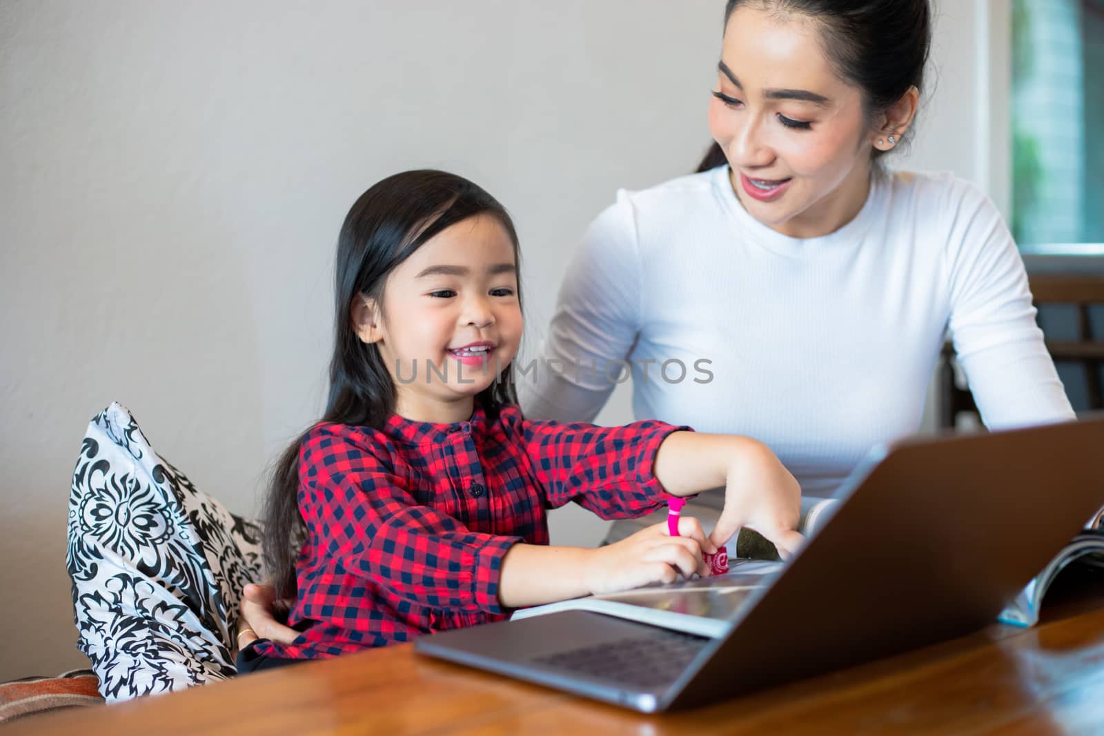 Asian mothers are teaching their daughters to read a book and use notebooks and technology for online learning during school holidays at home. Educational concepts and activities of the family