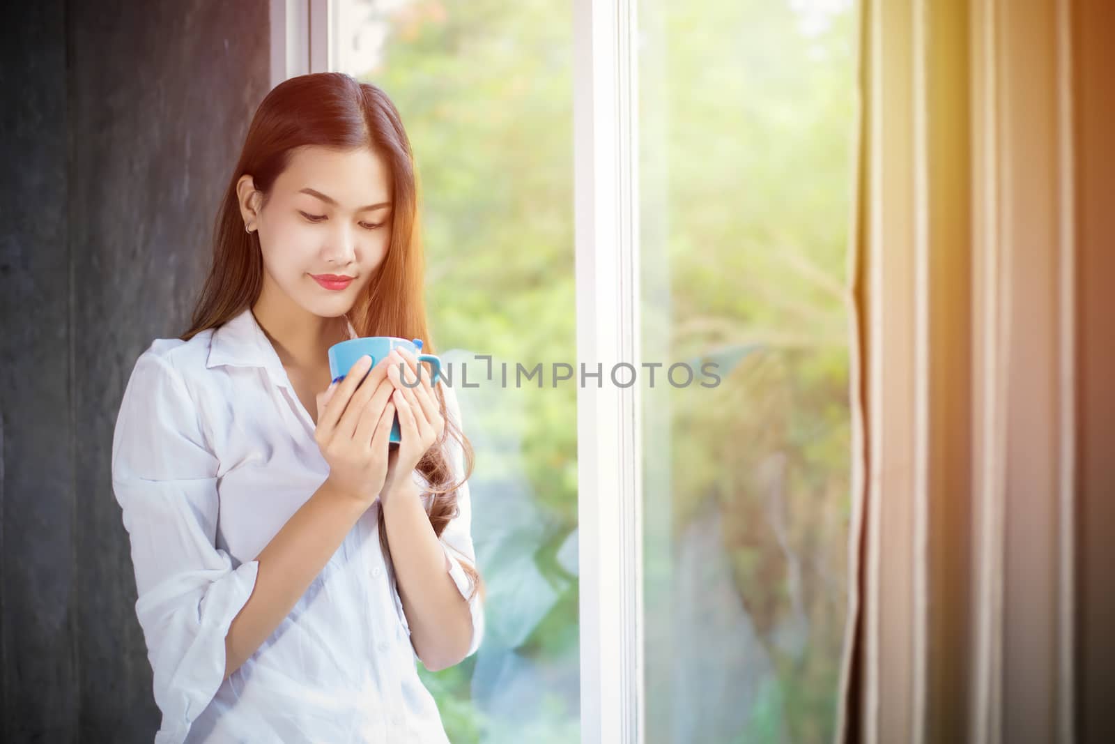 An Asian woman stood holding a coffee cup and opened the curtain on the window after waking up in the morning of the day at home