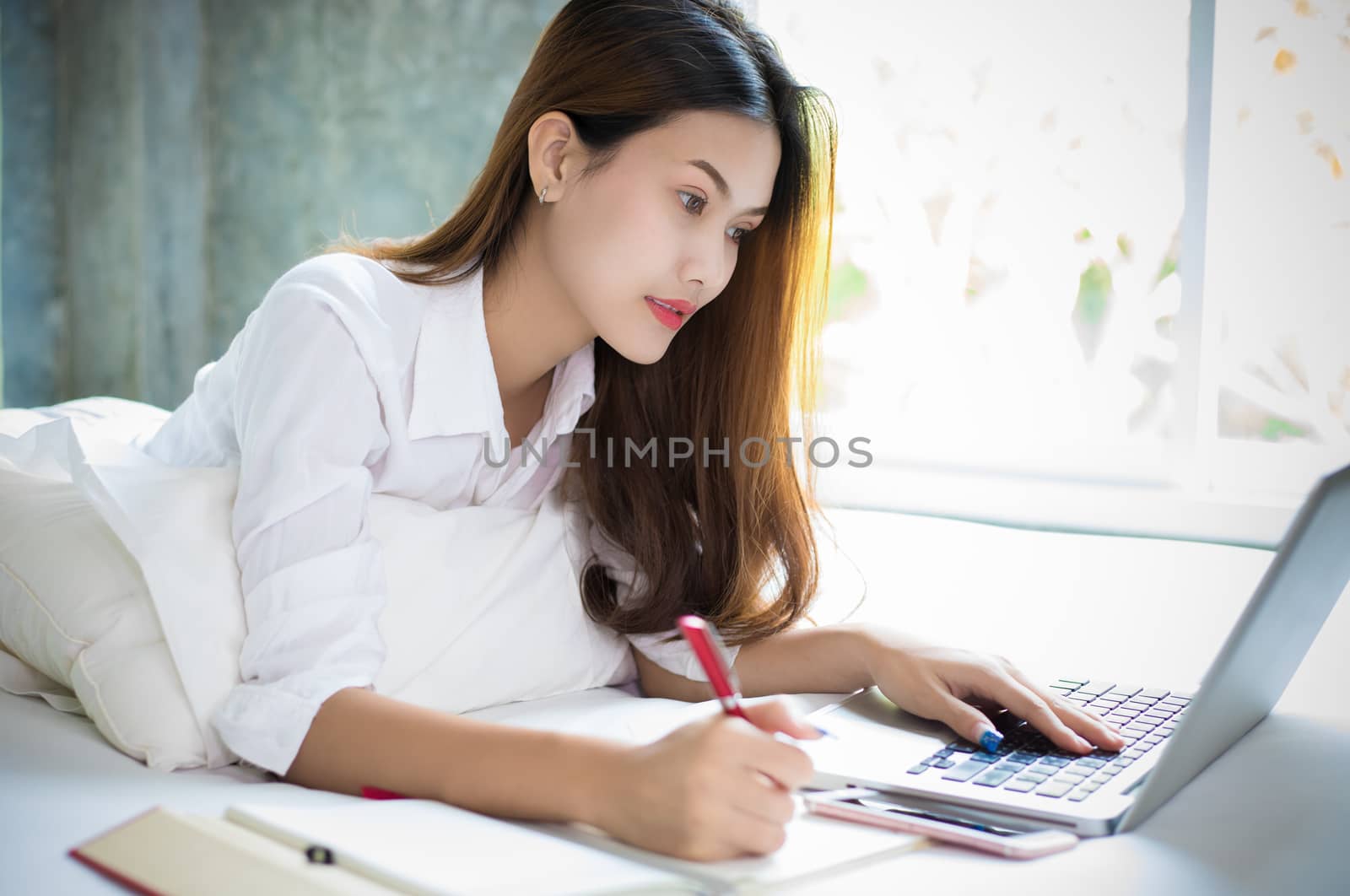 Asian woman is using a pen to write notes in her notebook and she uses a notebook to work on her bed at home.