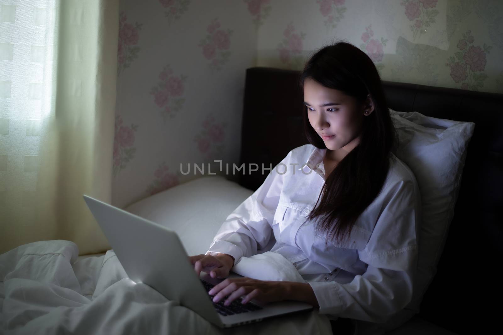 Asian woman using a notebook to work on night at the bed.She smiles and enjoys working at home.