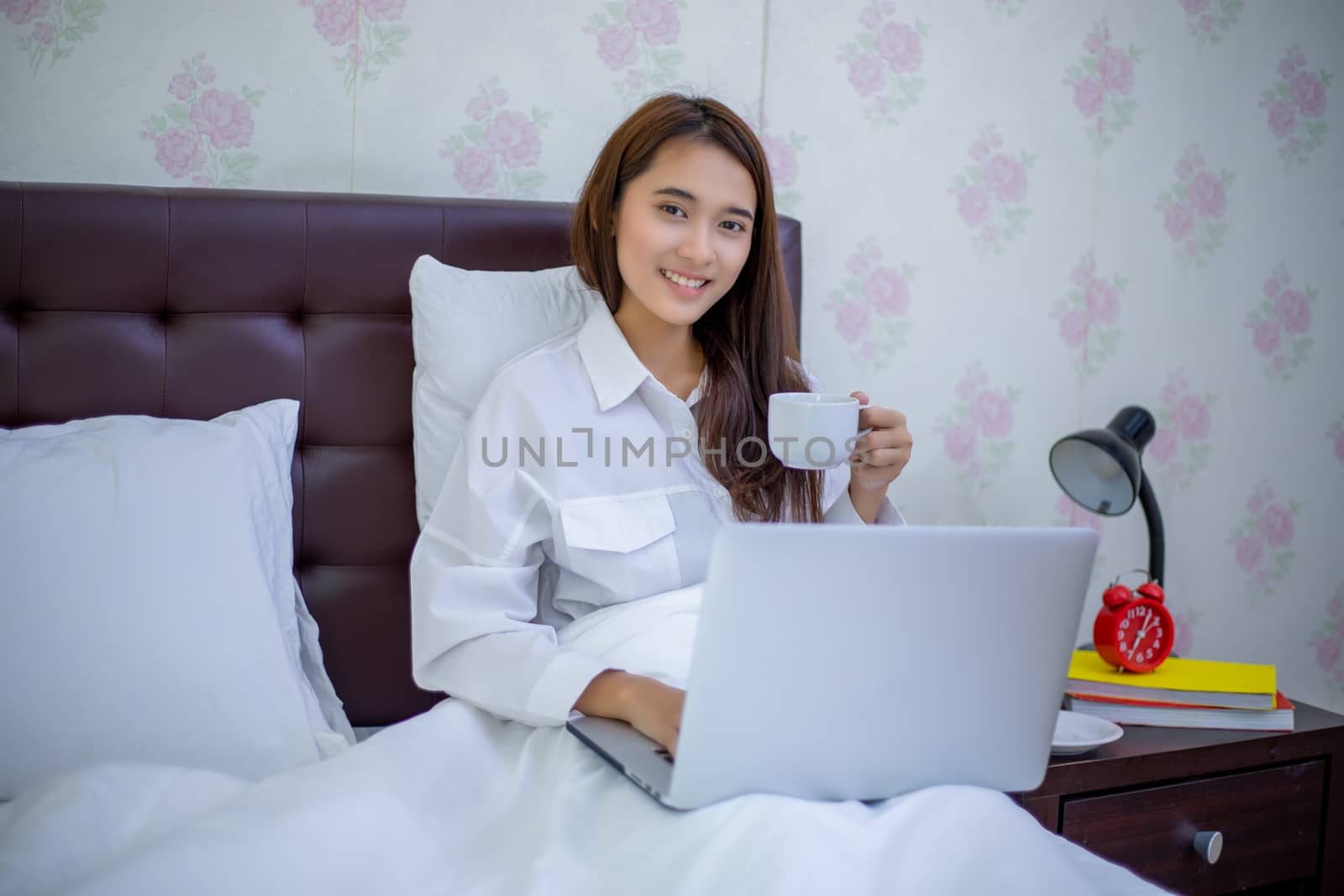 Asian woman is drinking a cup of coffee and using a notebook to work on the bed.She smiles and enjoys working at home.