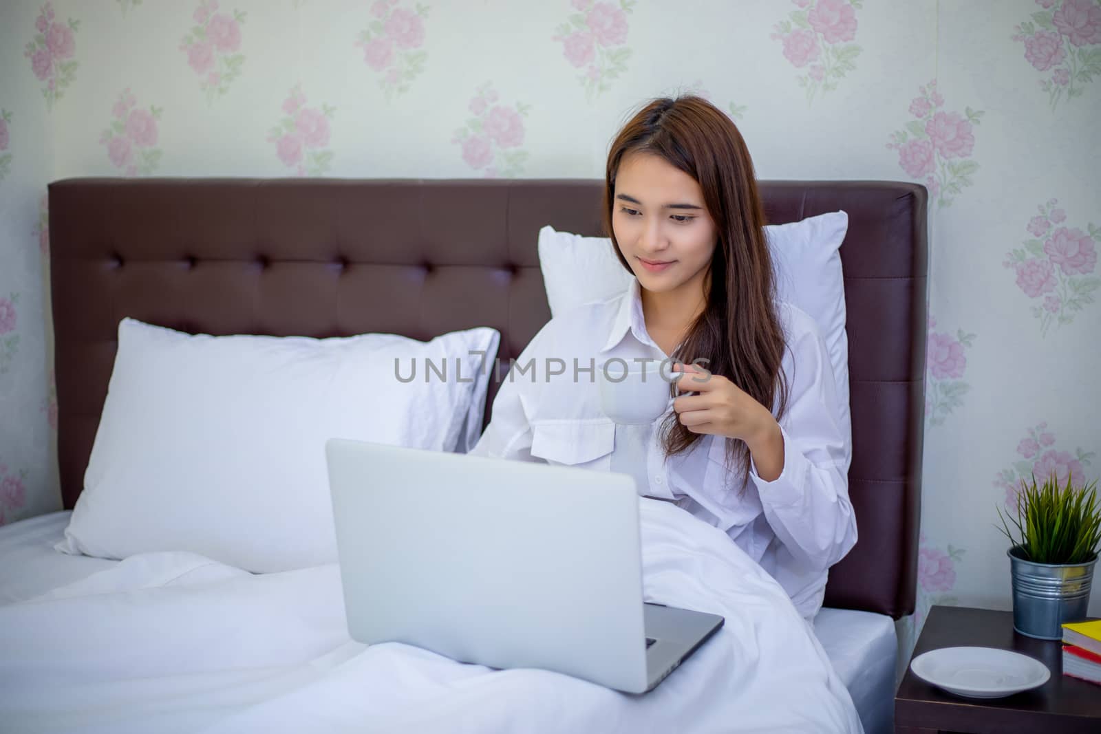 Asian woman is drinking a cup of coffee and using a notebook to work on the bed.She smiles and enjoys working at home.