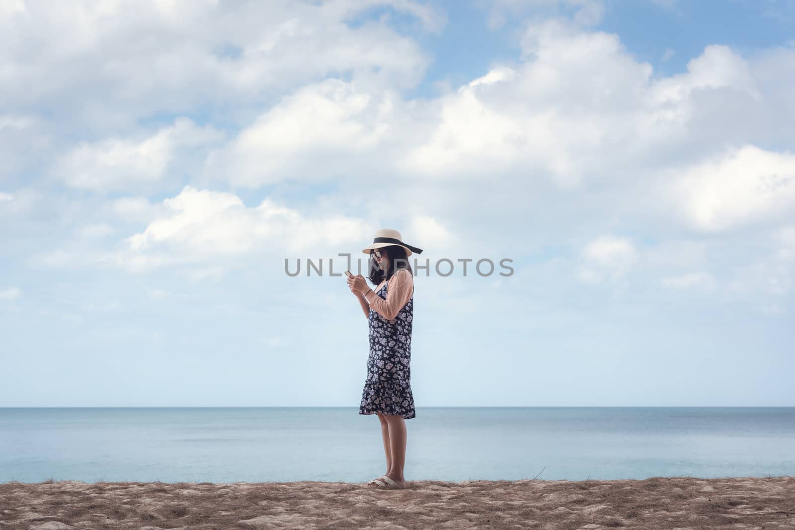 Portrait of Asian Woman is Using Mobile Phone Against Blue Sky and Sand Beach, Beautiful Woman is Having Fun and Enjoying With Her Phone While Summer Vacation on The Beach. Relaxation Lifestyles
