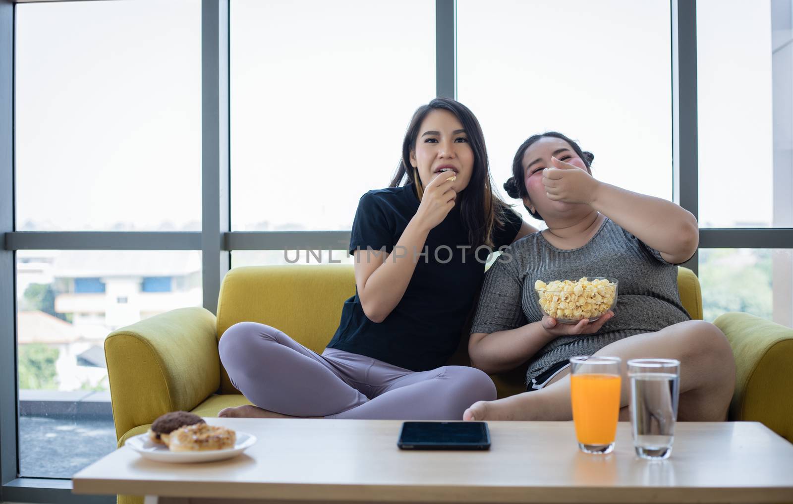 Overweight woman and asian girl enjoy eating food on sofa at home