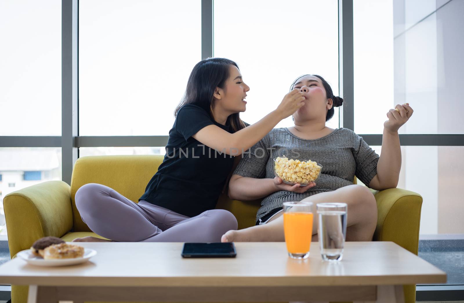 Overweight woman and asian girl enjoy eating food on sofa at home