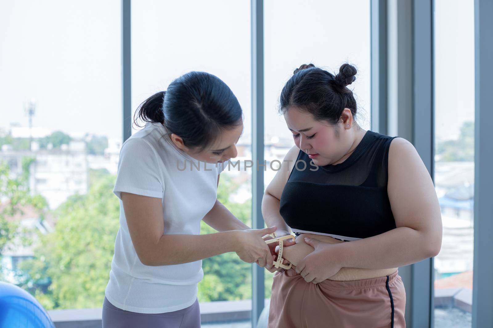 Young female trainer measuring fat layer of overweight woman with caliper at fitness