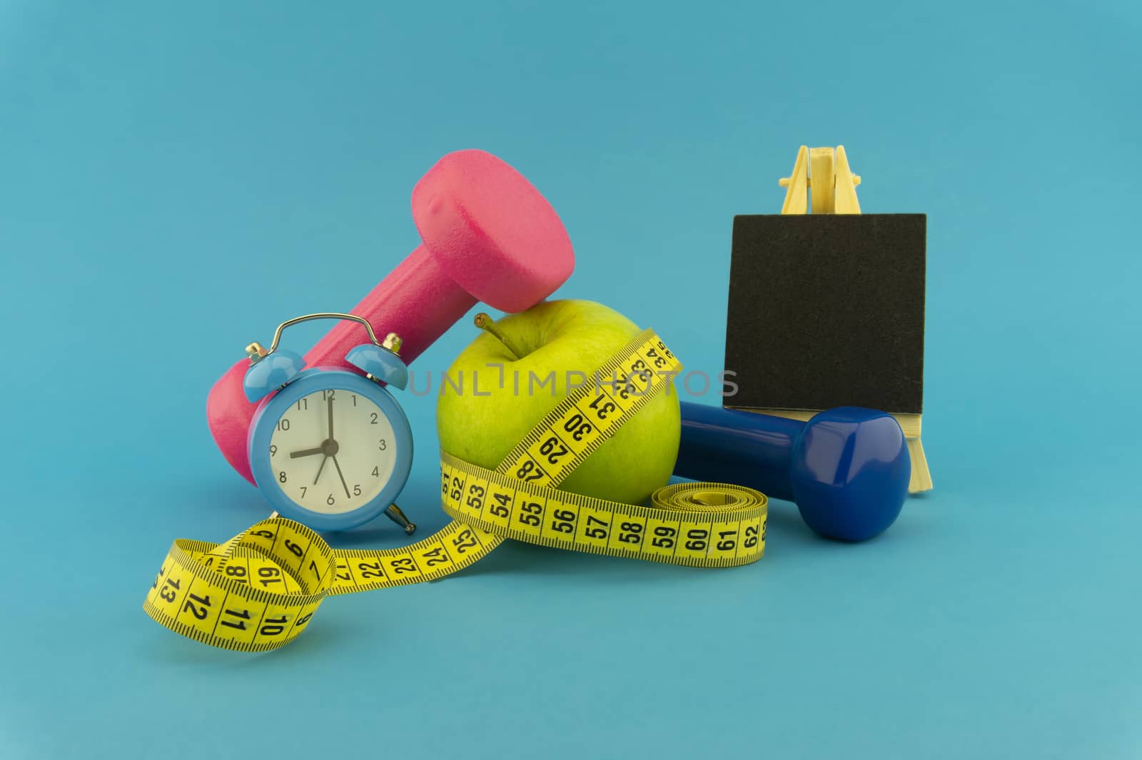 Starting healthy lifestyle concept. Still life with pink and blue dumbbells, measuring tape, apple, chalkboard and alarm clock on blue background