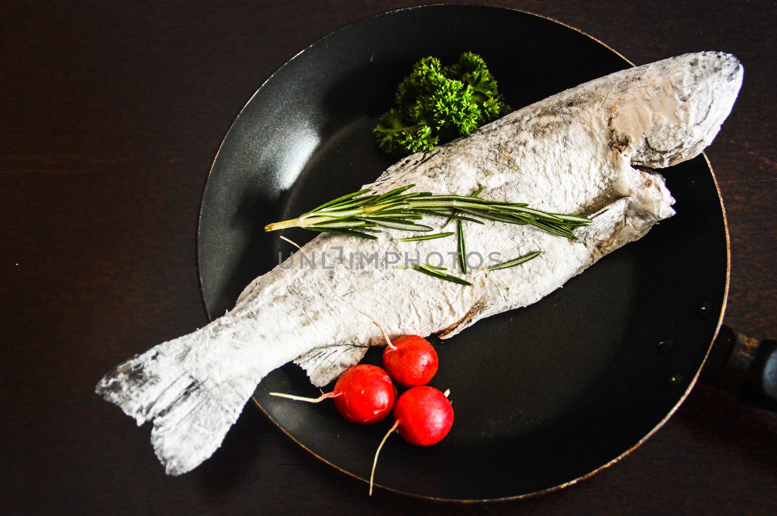 Top view of a trout fish covered in flour topped with rosemary and parsley in an iron skillet by Sonnet15