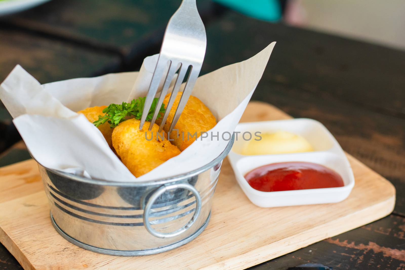 Delicious deep fried chicken nuggets on white paper in metal bucket on wooden tray and rustic table with blurry ketchup and mayonnaise. Unhealthy food concept. Eatting with fork.