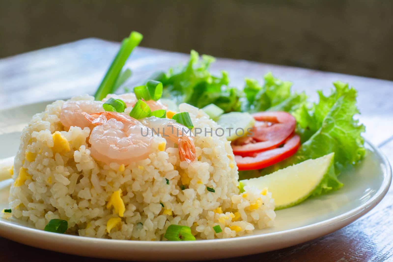 Close up fried rice with shrimp, egg and green onion on white plate serve with blurry cucumber, red tomato, lettuce in cafe and restaurant.