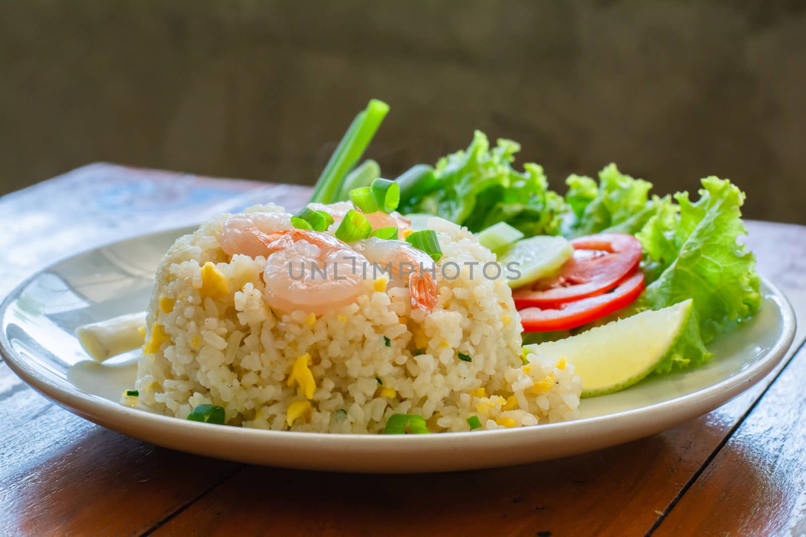 Close up fried rice with shrimp, egg and green onion on white plate serve with blurry cucumber, red tomato, lettuce in cafe and restaurant.