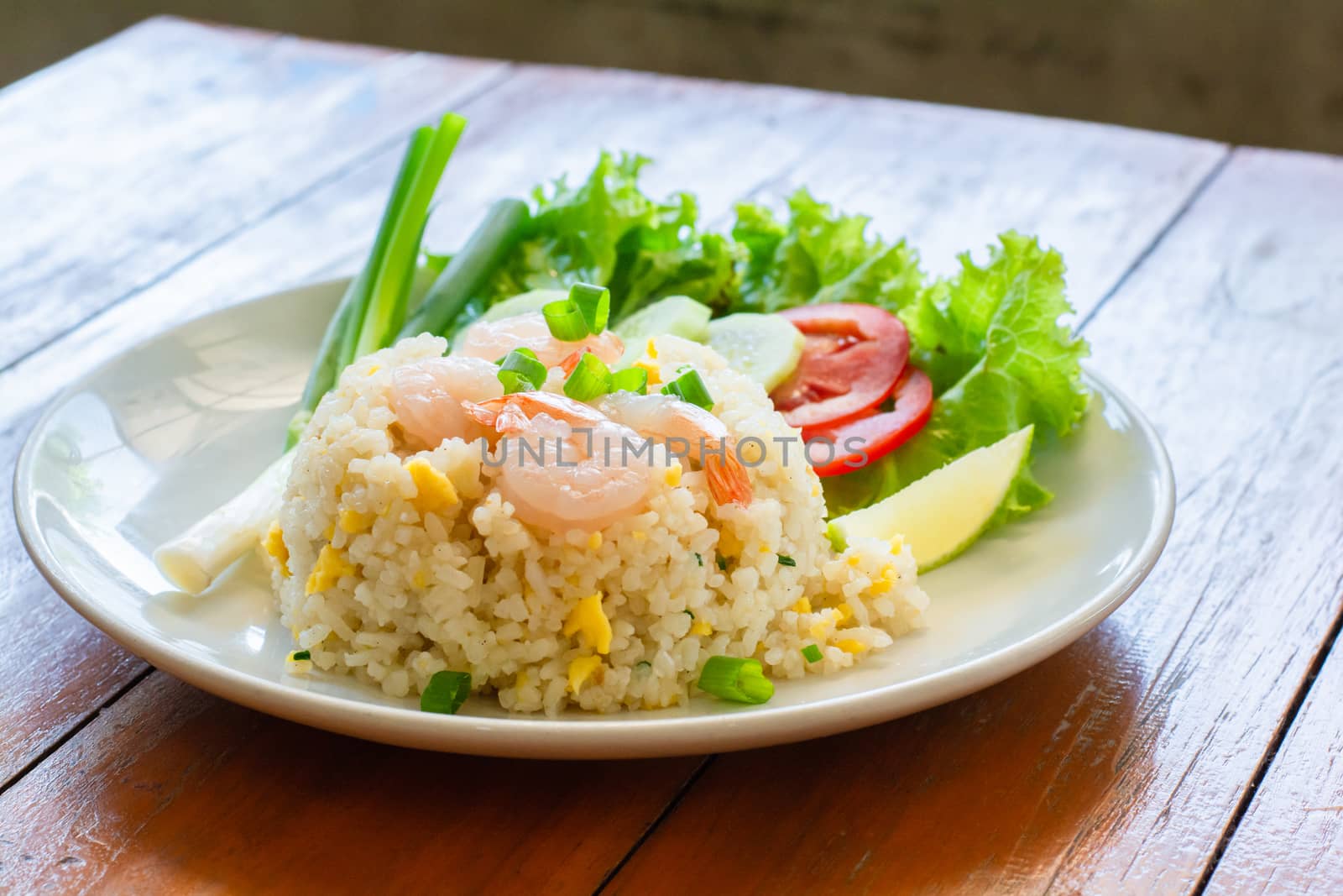 Close up fried rice with shrimp, egg and green onion on white plate serve with blurry cucumber, red tomato, lettuce in cafe and restaurant.