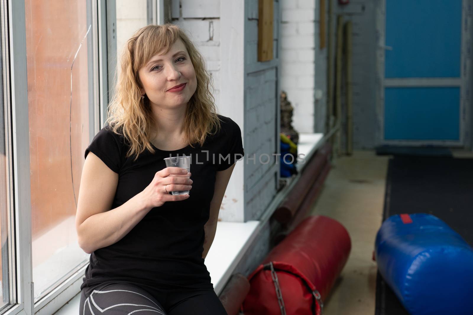 30-year-old woman with blond hair sits on a window sill against a window after a workout with a glass of water