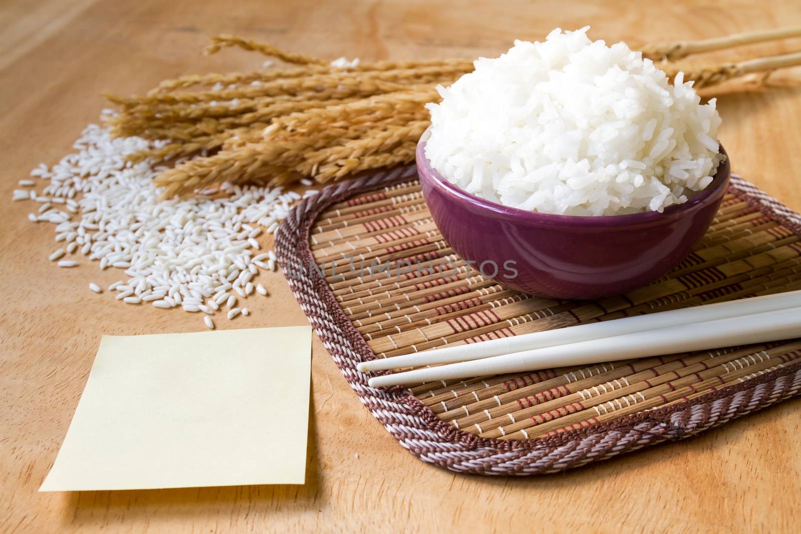 Cooked rice in bowl with raw rice grain ,dry rice plant and empty paper on wooden table background. by ronnarong