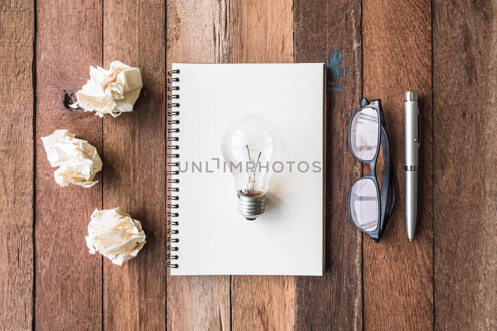 Top view of notebook with crumpled paper balls, pen, glasses and light bulb on wooden table background.