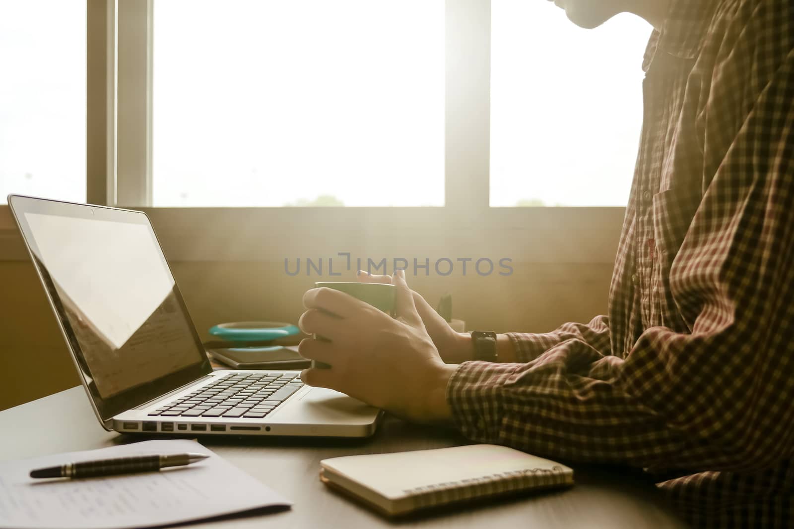 A man holding a cup of coffee and working on laptop on his office desk,toned with sunlight.