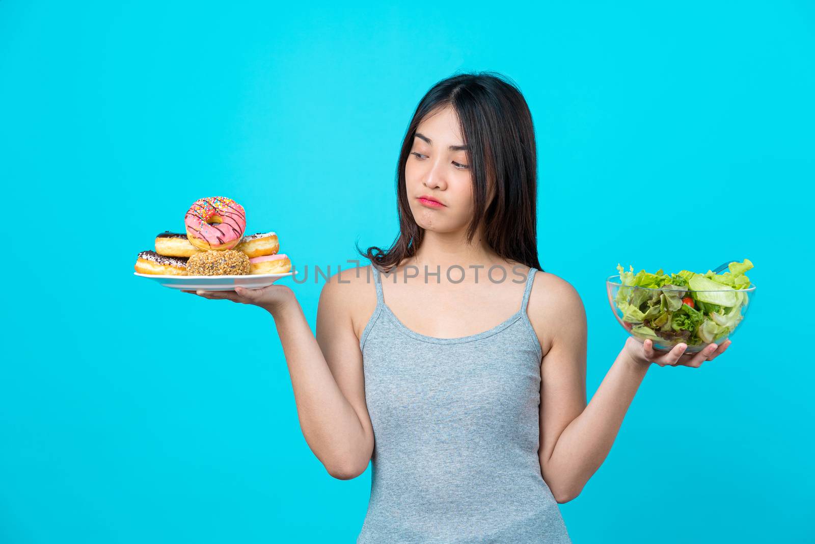 Attractive Asian young woman holding and choosing between disk of donuts or vegetable salad in glasses bowl on isolated blue color background, weight loss and avoid junk food for dieting and Healthy